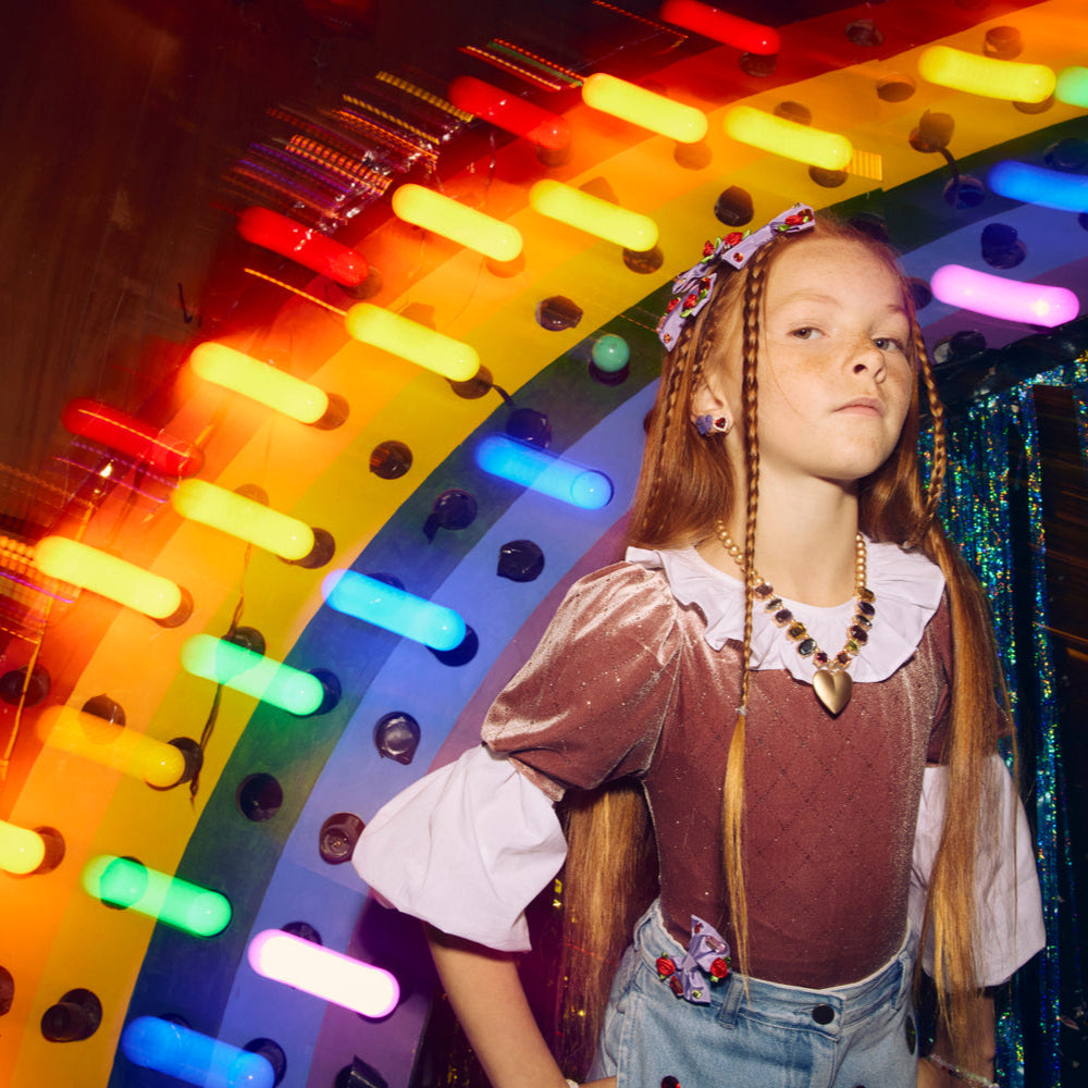 Child standing in front of a colorful neon rainbow sign. she is wearing lots of hair accessories and a fun chunky necklace.