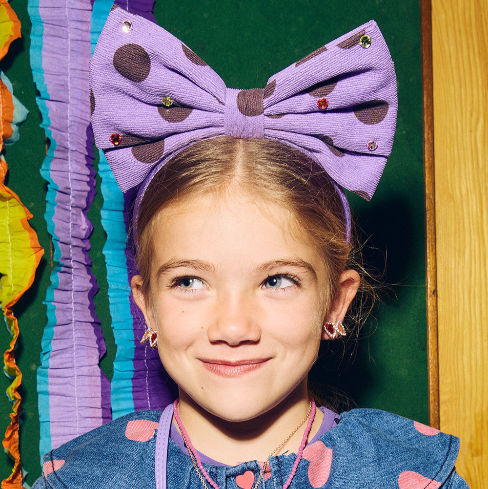 Young girl wearing a large purple bow headband with polka dots, smiling against a colorful background.